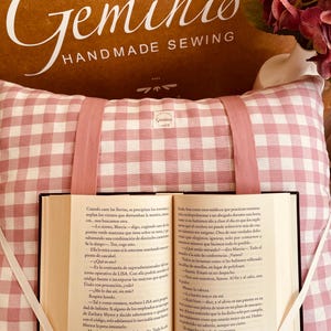 Può includere: Un cuscino rosa e bianco a quadretti con un libro e un segnalibro a nastro. Il libro è aperto, rivelando il testo. Il cuscino ha un cinturino rosa e le parole "Géminis HANDMADE SEWING" sono visibili.