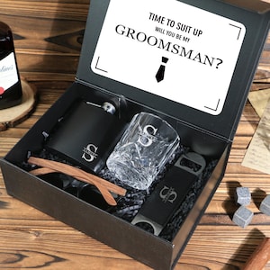 Puede incluir: Una caja de regalo negra que contiene una petaca, un vaso de whisky, un abrebotellas y gafas de sol. La petaca y el vaso están grabados con la letra "S". La tapa de la caja tiene el texto "TIME TO SUIT UP WILL YOU BE MY GROOMSMAN?"