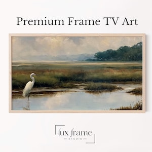 May include: A landscape painting in a light wood frame titled Premium Frame TV Art. The artwork depicts a white egret standing in shallow water within a marshy wetland, with green grasses, distant trees, and a cloudy sky. The logo Lux Frame Studio 2022 is below.