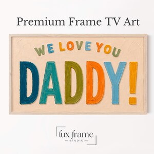 Puede incluir: Un marco rectangular de madera clara que contiene arte texturizado con el texto WE LOVE YOU DADDY! en letras multicolores en relieve, estilo pincelada. El fondo es de un color beige claro sólido con una textura sutil e irregular.