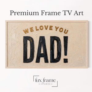 May include: A framed art piece with the words "WE LOVE YOU DAD!" in gold and black. The art has a textured, off-white background and a light wood frame. The words are in a bold, stylized font.