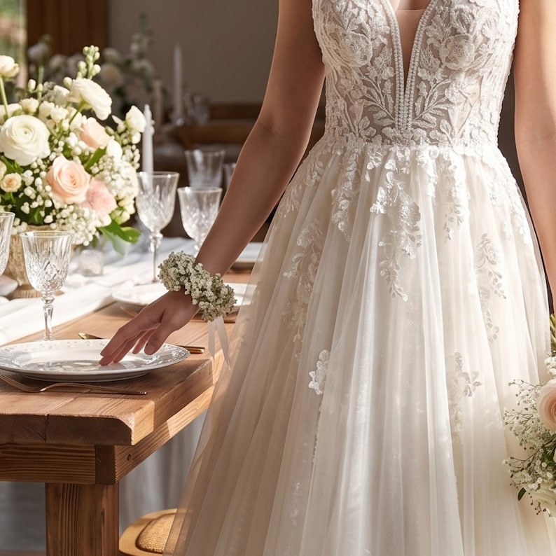 May include: A white wedding dress with a deep V-neckline and floral lace detailing. The dress has a flowing tulle skirt. A floral wrist corsage and a bouquet of flowers are also visible. The table is set with white plates, crystal glasses, and a floral centerpiece.