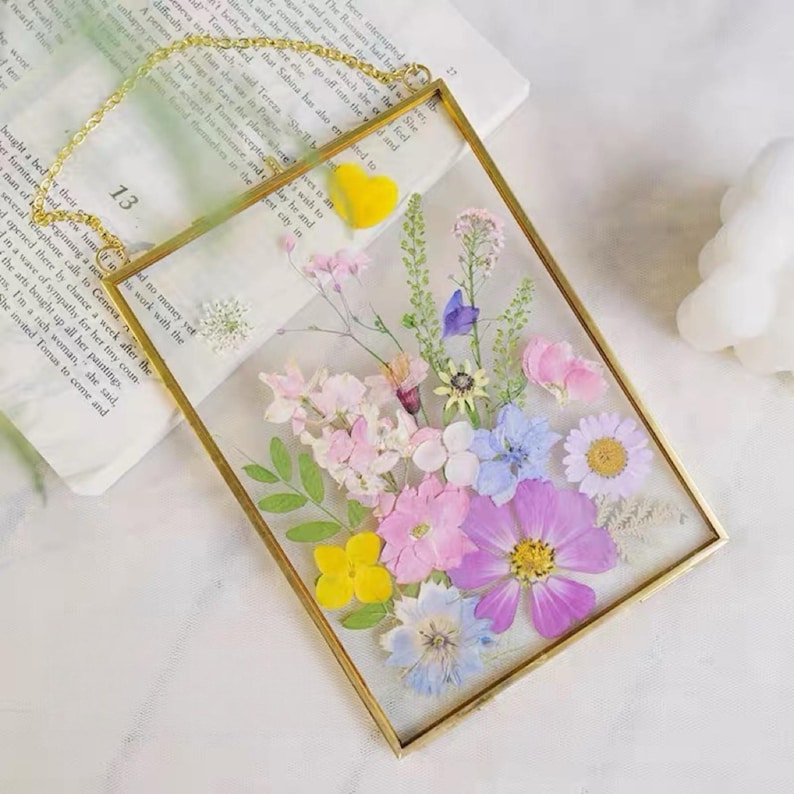 May include: A rectangular gold-framed pressed flower art piece. The artwork features a vibrant arrangement of dried flowers in shades of pink, purple, yellow, and blue. A gold chain is attached for hanging.