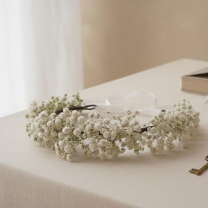 May include: A delicate floral crown featuring small white flowers. The crown is made of a dark brown wire base and adorned with clusters of tiny white blossoms. A white ribbon is attached for tying. The crown is a beautiful accessory for special occasions.
