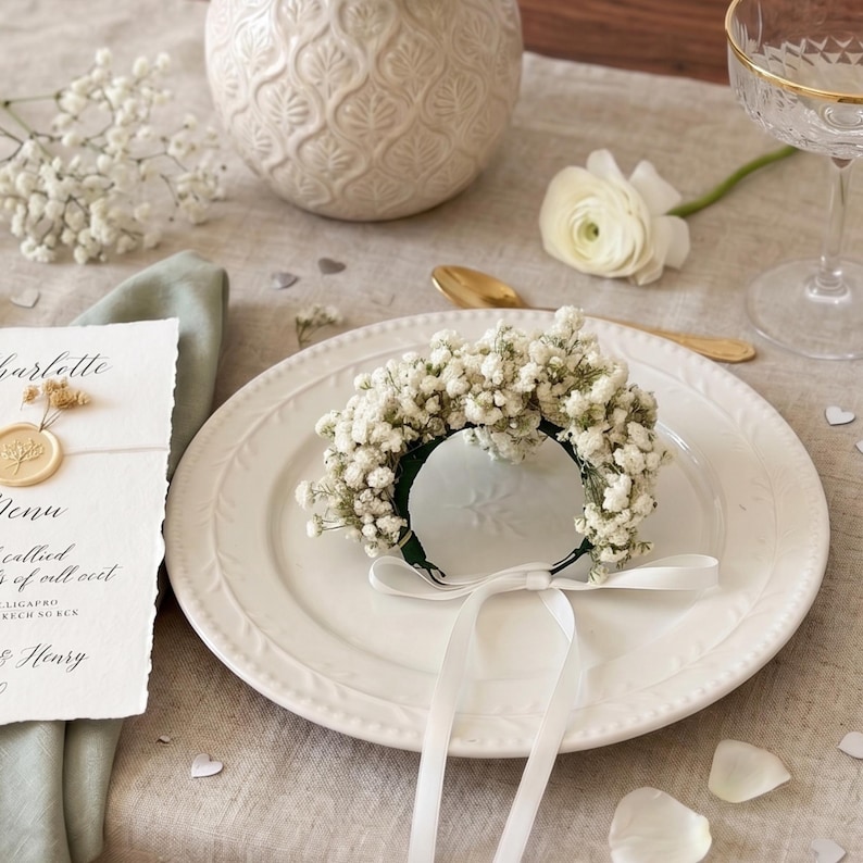 May include: A white plate with a floral napkin ring made of baby's breath and a white ribbon. A menu card with a gold wax seal and a white flower are also visible. The setting is on a linen tablecloth.