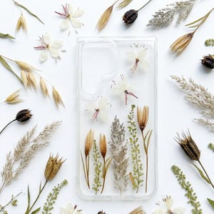 May include: A clear phone case with pressed flowers and foliage. The case features a variety of dried plants, including white flowers, brown seed pods, and green stems. The arrangement is set against a white background, creating a natural and elegant aesthetic.