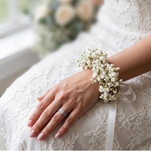 May include: A close-up of a hand wearing a diamond ring and a wrist corsage made of small white flowers. The wrist corsage is tied with a white ribbon. The person is wearing a white lace dress.