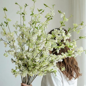 May include: A large bouquet of white artificial flowers with green stems and leaves. The flowers have a delicate, clustered appearance, and the stems are bundled together. The background is a plain white wall and a white curtain.