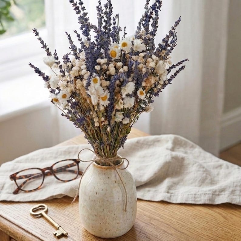 Op de afbeelding: Een boeket van gedroogde lavendel, madeliefjes en andere wilde bloemen in een gespikkelde keramische vaas vastgebonden met touw. De vaas staat op een houten tafel met een bril en een decoratieve sleutel. De bloemen hebben paarse, witte en gele tinten.