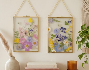 May include: Two rectangular gold-framed pressed flower art pieces hanging on a wall. The frames contain a variety of colorful dried flowers. A vase with pampas grass, books, a candle, and a plant are on a wooden shelf below.