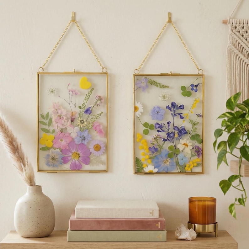 May include: Two rectangular gold-framed pressed flower art pieces hanging on a wall. The frames contain a variety of colorful dried flowers. A vase with pampas grass, books, a candle, and a plant are on a wooden shelf below.