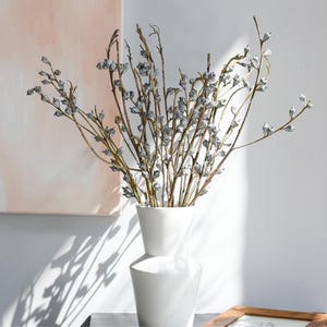 Op de afbeelding: Een witte keramische vaas met een geometrisch silhouet bevat een verzameling slanke, lichtbruine takken versierd met kleine, zilverkleurige, klokvormige zaaddoosjes. De takken waaieren uit vanuit de opening van de vaas.