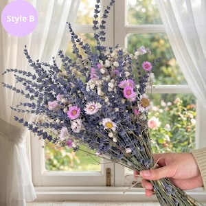 Op de afbeelding: Een boeket van gedroogde bloemen met lavendel, roze en witte bloemen. Het boeket is vastgebonden met touw en wordt vastgehouden voor een raam met witte gordijnen. De tekst "Style B" staat in een paarse cirkel.