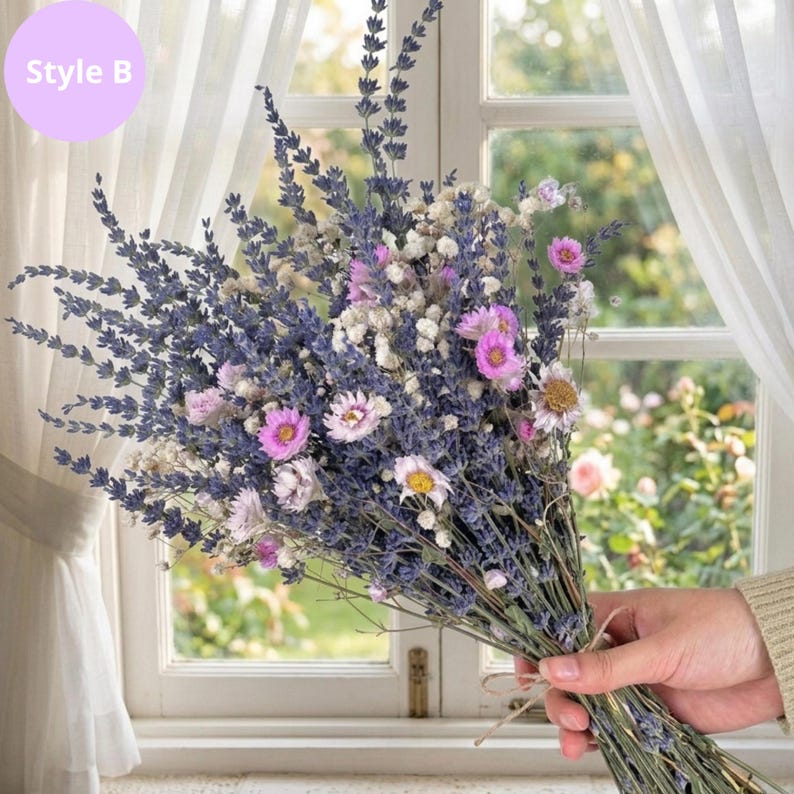 Op de afbeelding: Een boeket van gedroogde bloemen met lavendel, roze en witte bloemen. Het boeket is vastgebonden met touw en wordt vastgehouden voor een raam met witte gordijnen. De tekst "Style B" staat in een paarse cirkel.