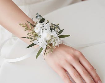 May include: A white floral wrist corsage featuring white flowers, green leaves, and sprigs of greenery. The corsage is tied with a white ribbon. The wrist corsage is a wedding or special occasion accessory.