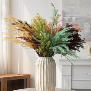 May include: A tall, cylindrical, off-white ceramic vase with vertical ribbed texture holds a large, dense arrangement of artificial feathery foliage in a multi-colored palette of yellow, sage green, light green, rust orange, and dark brown.