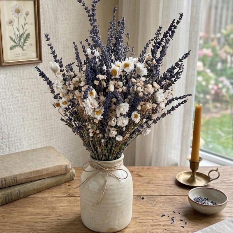 Op de afbeelding: Een boeket van gedroogde lavendel, witte madeliefjes en andere kleine witte bloemen in een cr&egrave;mekleurige keramische vaas, vastgebonden met touw. De vaas staat op een houten tafel met een kaars en een kleine kom met lavendelknopjes.