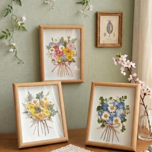 Op de afbeelding: Drie ingelijste bloemenkunstwerken met geperste bloemen. De boeketten bevatten een verscheidenheid aan kleuren, waaronder geel, roze en blauw. De frames zijn gemaakt van lichtgekleurd hout en worden op een houten oppervlak weergegeven.