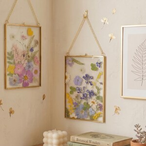 May include: Two rectangular gold-framed pressed flower art pieces hanging on a wall. The frames are filled with colorful dried flowers, including pink, purple, yellow, and blue blooms. The frames are suspended by gold chains.