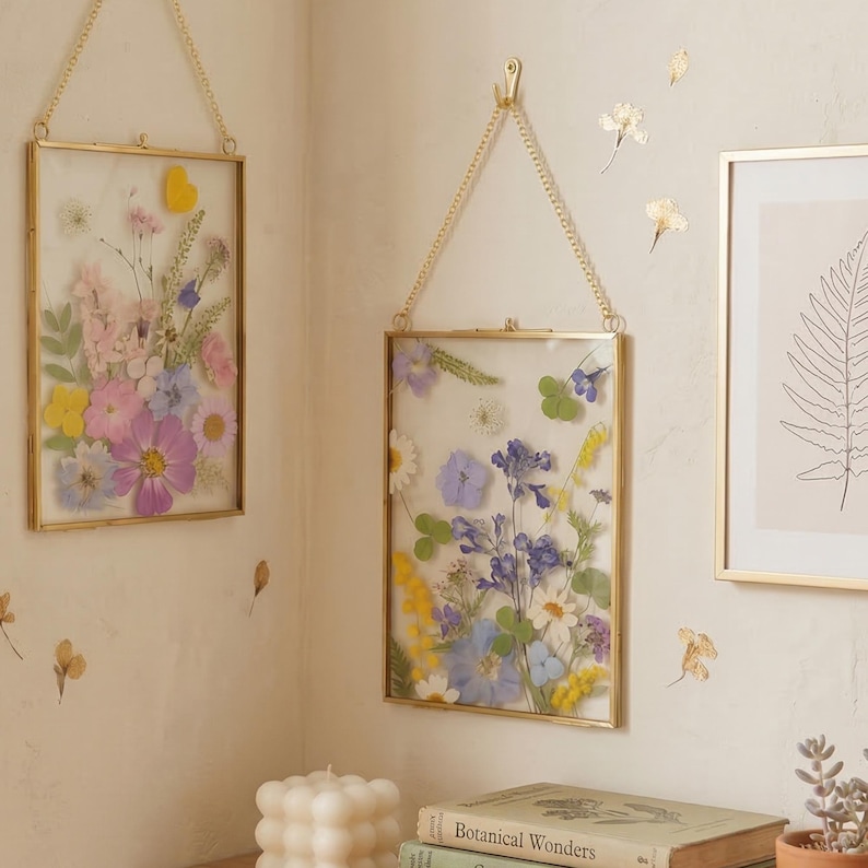 May include: Two rectangular gold-framed pressed flower art pieces hanging on a wall. The frames are filled with colorful dried flowers, including pink, purple, yellow, and blue blooms. The frames are suspended by gold chains.