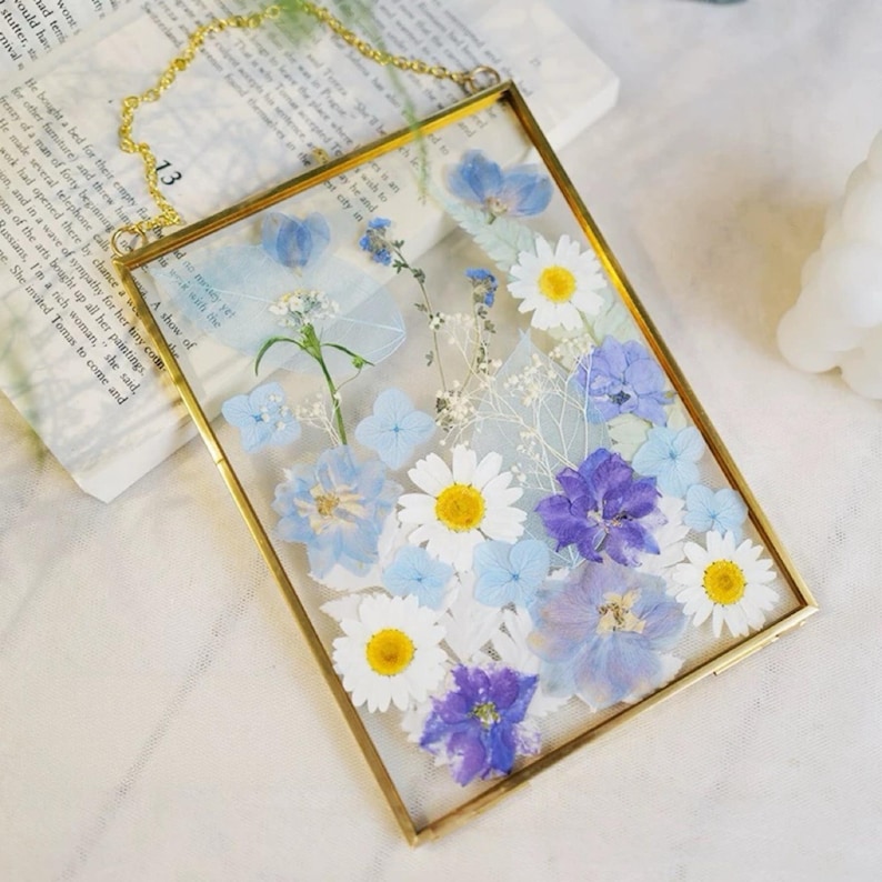 May include: A rectangular glass frame with a gold-colored metal border, filled with pressed flowers. The arrangement includes white daisies, blue hydrangeas, and purple blooms. A gold chain is attached for hanging.