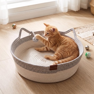 May include: A round, two-toned woven rope pet bed featuring a cream-colored base and a gray upper rim with integrated cat-ear-shaped handles. A small, dangling gray rope ball toy is attached to one handle, and a brown leather-like brand tag is visible.