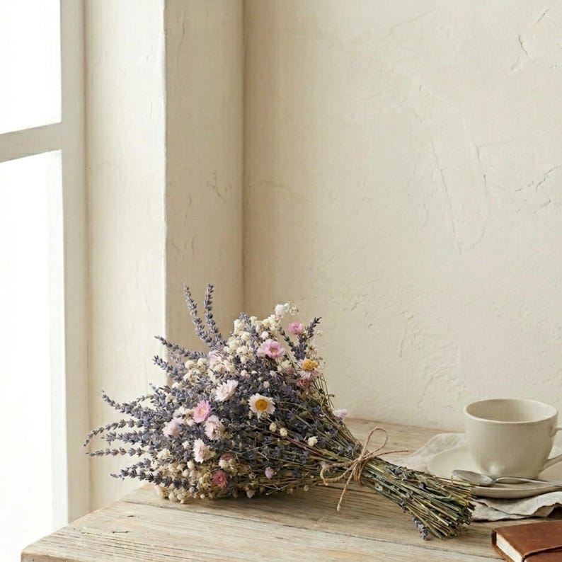 Op de afbeelding: Een boeket van gedroogde bloemen met lavendel, gipskruid en kleine roze en gele bloemen. Het boeket is vastgebonden met touw en ligt op een houten oppervlak. Een kop en schotel staan op de achtergrond.