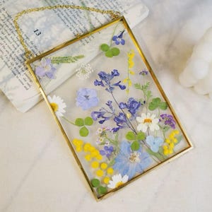 May include: A rectangular glass frame with a gold-colored metal border, filled with an assortment of dried flowers in shades of blue, yellow, white, and purple. The frame is suspended by a gold chain. The background includes a book with the number 13 visible.