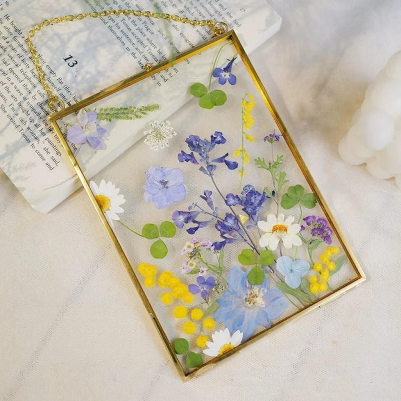 May include: A rectangular glass frame with a gold-colored metal border, filled with an assortment of dried flowers in shades of blue, yellow, white, and purple. The frame is suspended by a gold chain. The background includes a book with the number 13 visible.