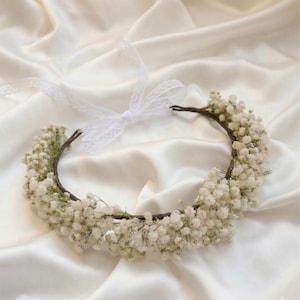 May include: A delicate floral crown featuring a circular base adorned with clusters of small, white flowers. A white lace ribbon is tied at the top, adding a touch of elegance. The crown is set against a backdrop of soft, white fabric, creating a romantic and ethereal feel.