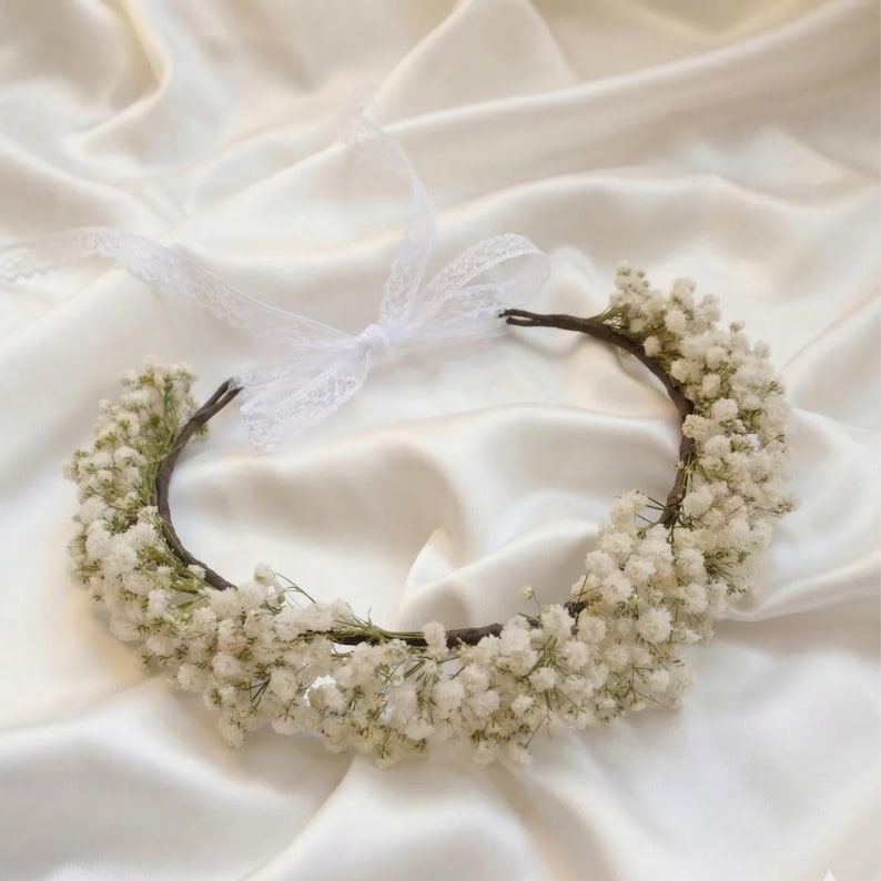 May include: A delicate floral crown featuring a circular base adorned with clusters of small, white flowers. A white lace ribbon is tied at the top, adding a touch of elegance. The crown is set against a backdrop of soft, white fabric, creating a romantic and ethereal feel.