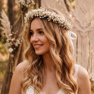 May include: A woman with long, wavy blonde hair wearing a delicate floral crown made of small white flowers. She is smiling, and the background includes natural elements like trees and decorative floral arrangements, suggesting a wedding or outdoor event.