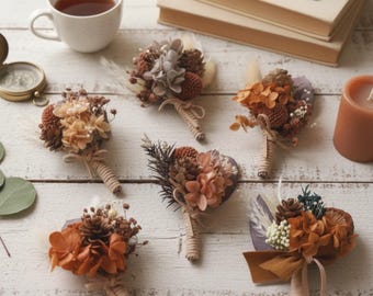 May include: An assortment of six dried flower boutonnieres in shades of brown, orange, and cream, arranged on a white wooden surface. Each boutonniere features a unique combination of dried flowers, pinecones, and greenery, wrapped with twine and ribbon. A cup of tea and books are in the background.