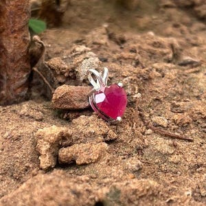 May include: A heart-shaped ruby pendant with a silver setting. The vibrant red gemstone is cut to a smooth, rounded heart shape. The pendant is displayed on a natural, earthy background, highlighting the jewelry's colour and design.