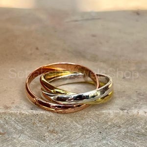 Peut inclure: Trois anneaux entrelacés en or rose, argent et or jaune. Les anneaux sont lisses et polis, reposant sur une surface texturée brun clair. Les anneaux sont un bijou.