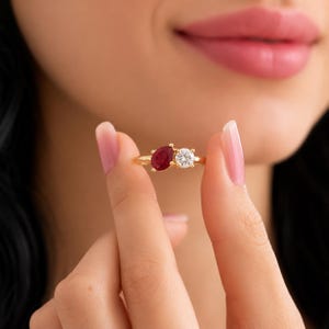 Può includere: Un anello in oro con due pietre incastonate fianco a fianco su una fascia sottile. Una pietra è una gemma rosso scuro a forma di pera, l'altra è una gemma trasparente a taglio rotondo. Entrambe sono fissate da piccole griffe in metallo dorato.