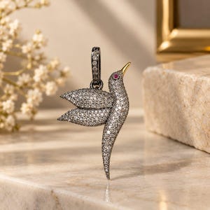 Può includere: Un ciondolo a forma di colibrì in metallo scuro, interamente tempestato di piccole pietre trasparenti incastonate a pavé. L'uccello presenta una piccola pietra rossa per l'occhio e un becco liscio color oro. È presente una contromaglia abbinata.