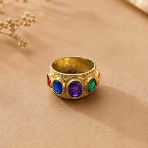 May include: A gold-toned metal ring featuring a wide, textured band set with multiple oval-shaped faceted gemstones in vibrant colours including red, blue, purple, green, and orange, arranged horizontally around the circumference of the ring.