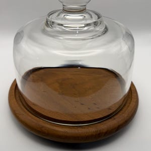 May include: A clear glass cloche with a rounded top and a small knob sits atop a round, wooden base. The base has a slightly raised edge and a rich, brown color. The cloche is designed to cover and protect items.