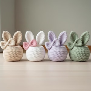 Może przedstawiać: Cztery szydełkowe pojemniki w kształcie królika w pastelowych kolorach: beżowym, białym, lawendowym i zielonym. Każdy ma kokardę i wyprostowane uszy. Pojemniki stoją na jasnej drewnianej powierzchni.