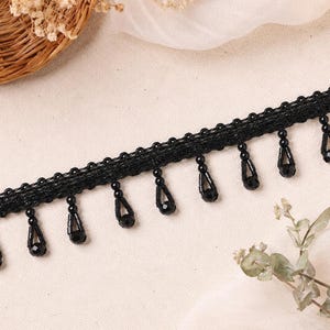 May include: Black beaded fringe trim with teardrop-shaped beads. The trim features a row of small, round beads along the top edge. This decorative trim is suitable for various craft and sewing projects.