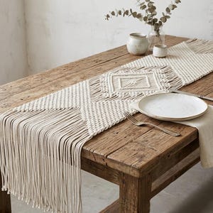 May include: A cream-coloured macrame table runner featuring intricate knotted geometric patterns and long, flowing fringe ends. The runner is laid out flat, showcasing a central diamond motif and textured cord construction across its length.