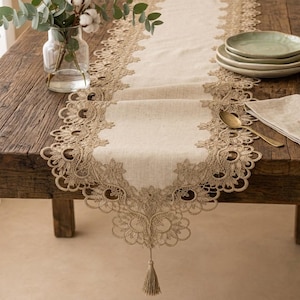 Pode incluir: Um caminho de mesa de linho bege com bordas de renda em tom bronze com padrões florais e uma borla decorativa na extremidade. O caminho está centrado numa mesa de madeira rústica, exibindo um design de bordado detalhado nas bordas.