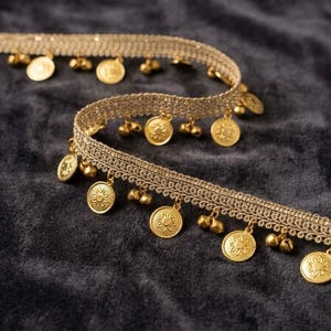 Op de afbeelding: Een decoratieve bies bestaande uit een geweven beige stoffen band, versierd met een herhalend patroon van kleine, bungelende goudkleurige metalen munten en piepkleine belletjes. De metalen elementen zijn gelijkmatig langs de onderrand geplaatst.