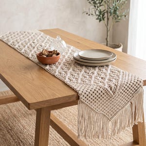 Puede incluir: Un camino de mesa de macramé color crema con un patrón tejido en forma de diamante y bordes con flecos largos. El camino está extendido sobre una mesa de madera, mostrando detalles de cuerdas anudadas y un acabado texturizado en algodón crudo.