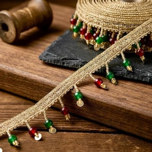 Può includere: Una bobina di nastro metallico dorato con una frangia decorativa di perline rosse e verdi alternate. Il nastro è realizzato con una rete dorata a trama aperta, con ogni perlina che pende da un piccolo anello di filo dorato sul bordo.