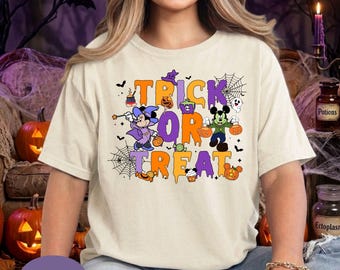 May include: An ivory t-shirt featuring a colorful Halloween graphic with the text "TRICK OR TREAT". The design includes cartoon characters in costumes, spider webs, pumpkins, and candy icons in shades of purple, orange, green, and black.