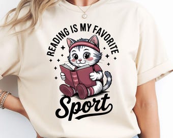May include: Cream-colored t-shirt featuring a cartoon cat reading a book. The cat wears a red headband and a matching outfit. The text "READING IS MY FAVORITE SPORT" is printed above and below the cat illustration.