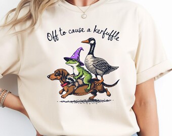 May include: Cream-colored t-shirt with a whimsical illustration. A dachshund dog, raccoon, frog in a wizard hat, and goose are stacked. The text "Off to cause a kerfuffle" is arched above the illustration in black.