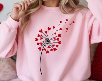 May include: A light pink sweatshirt featuring a black and red heart-shaped dandelion design. The dandelion's seeds are depicted as red hearts blowing in the wind. The sweatshirt is a solid light pink color.
